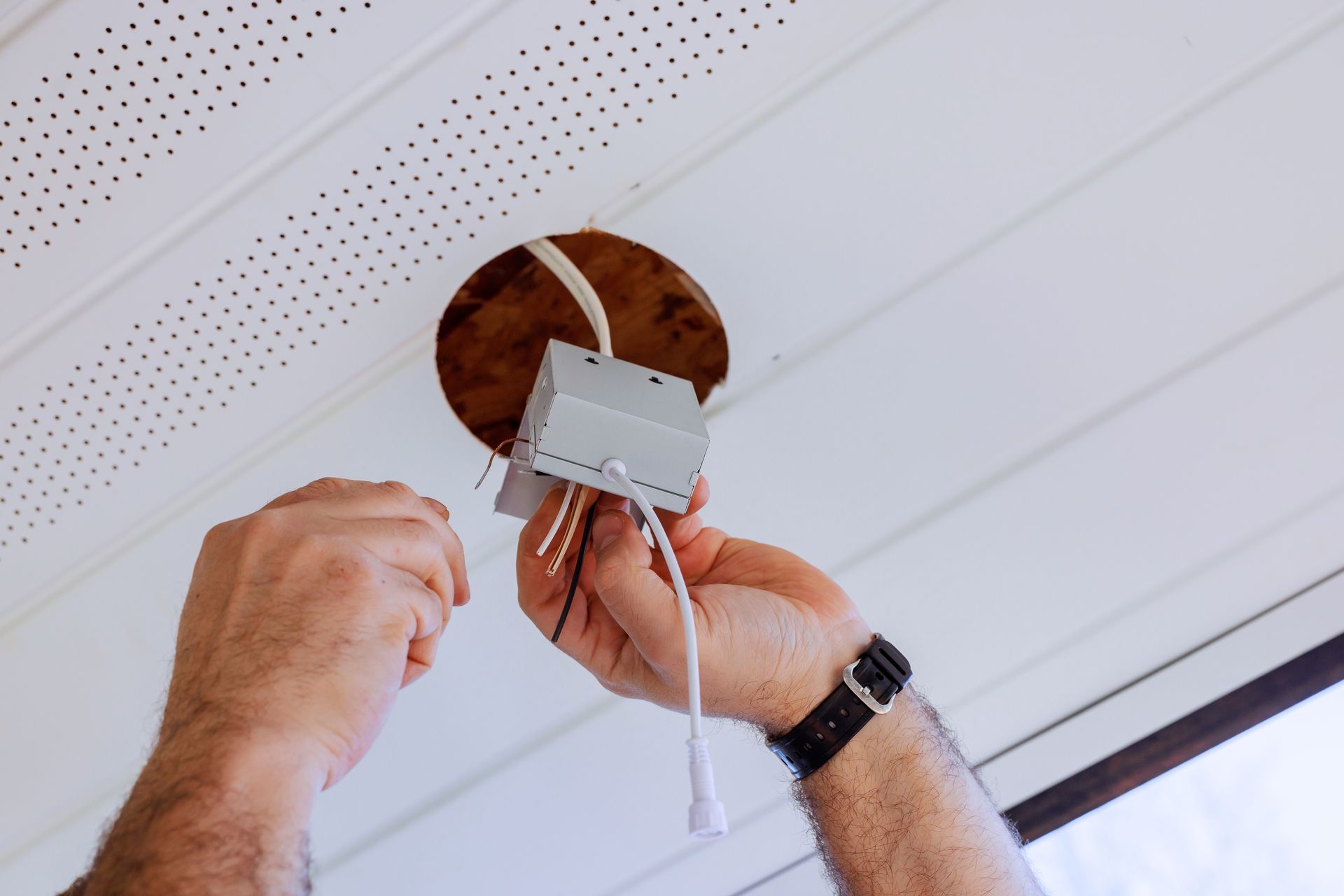 Hands installing a white electrical device in a ceiling opening with exposed wires