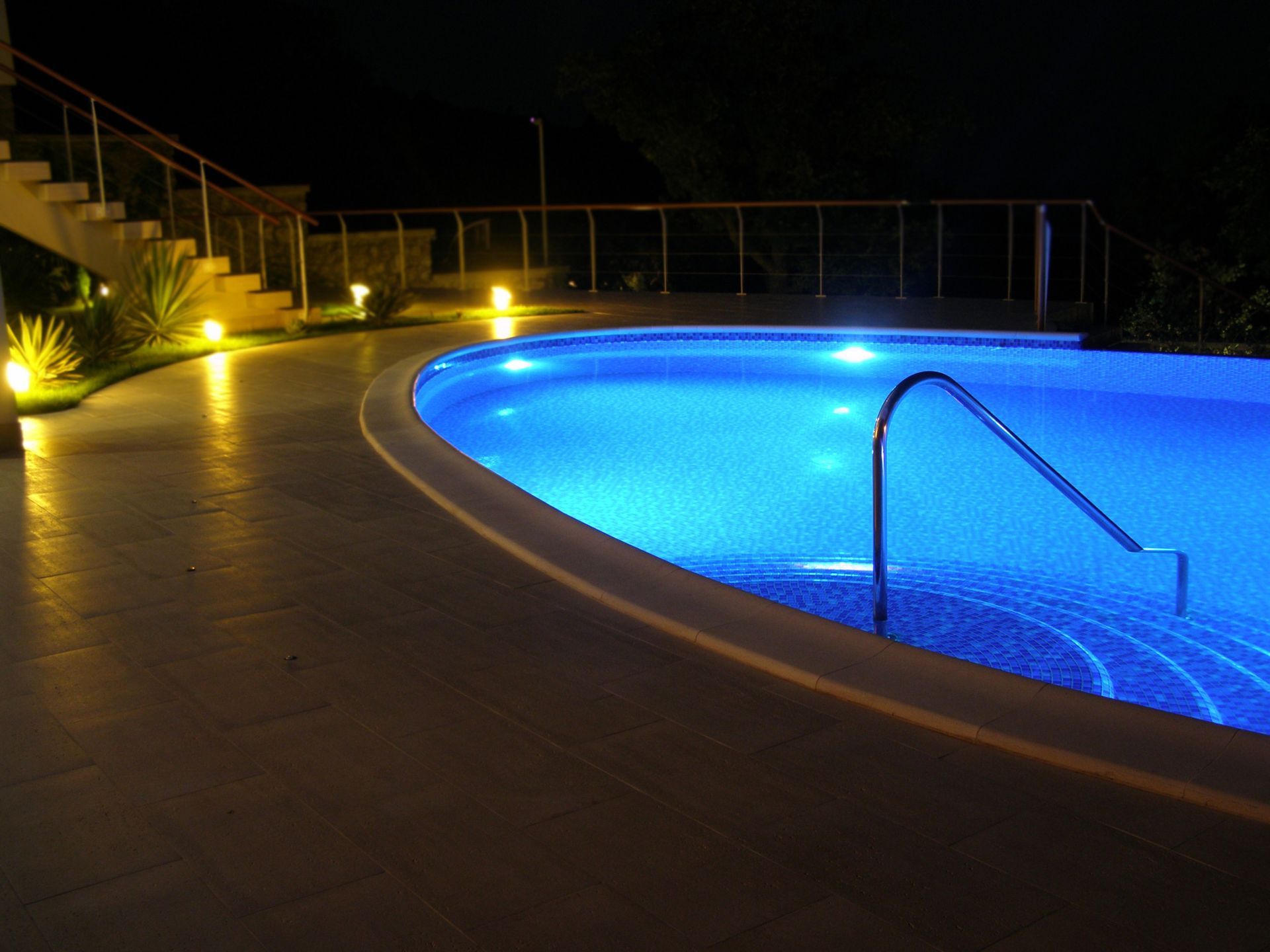 Night view of an illuminated blue swimming pool with a handrail. Pathway and stairs lit by warm lights.