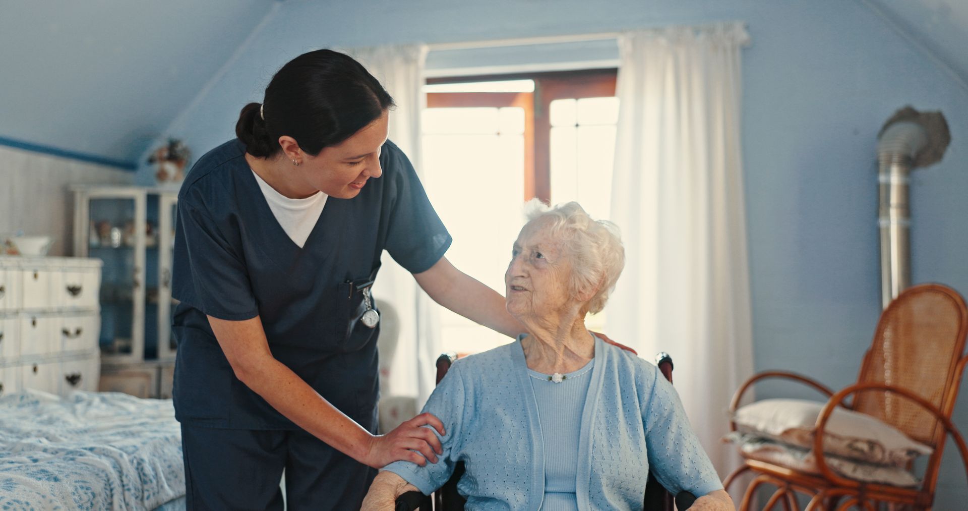 Caregiver in blue scrubs assisting an elderly person in a wheelchair indoors.