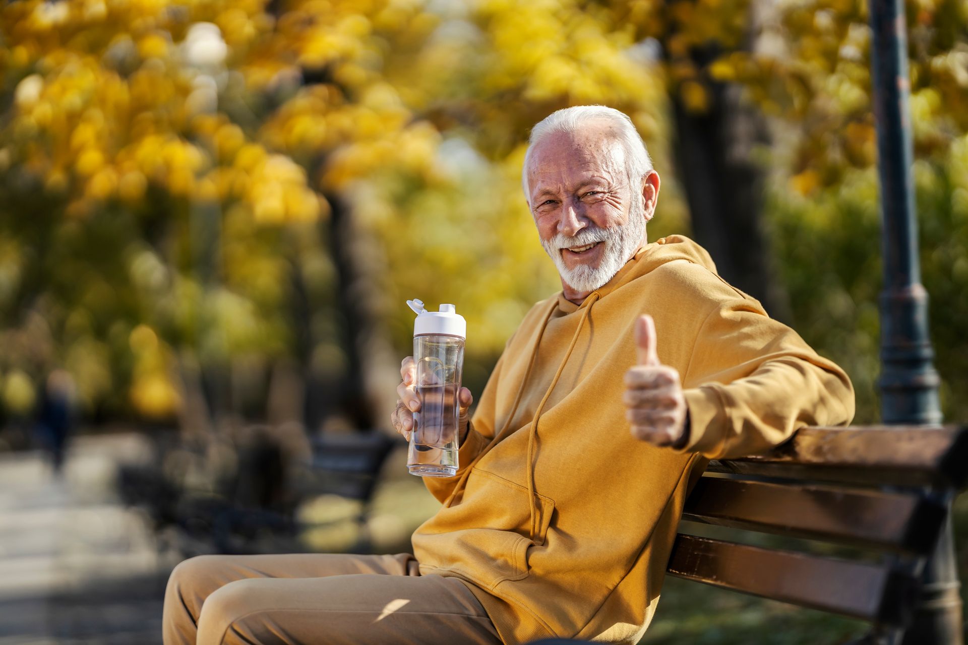 Elderly man in mustard hoodie sitting on a park bench, holding water bottle, giving thumbs up.