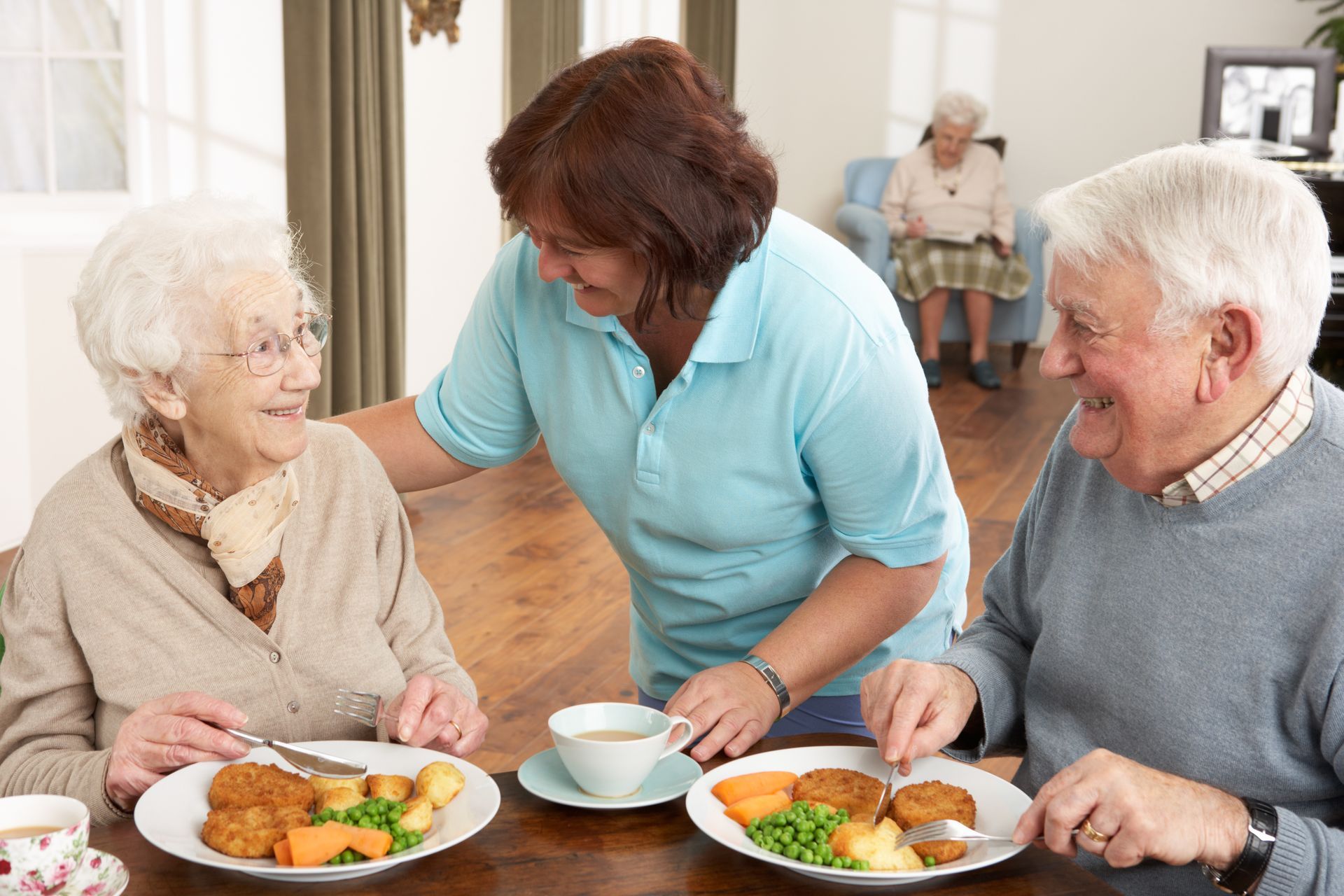A caregiver stands between two individuals dining at a table, smiling while helping them with their meals.