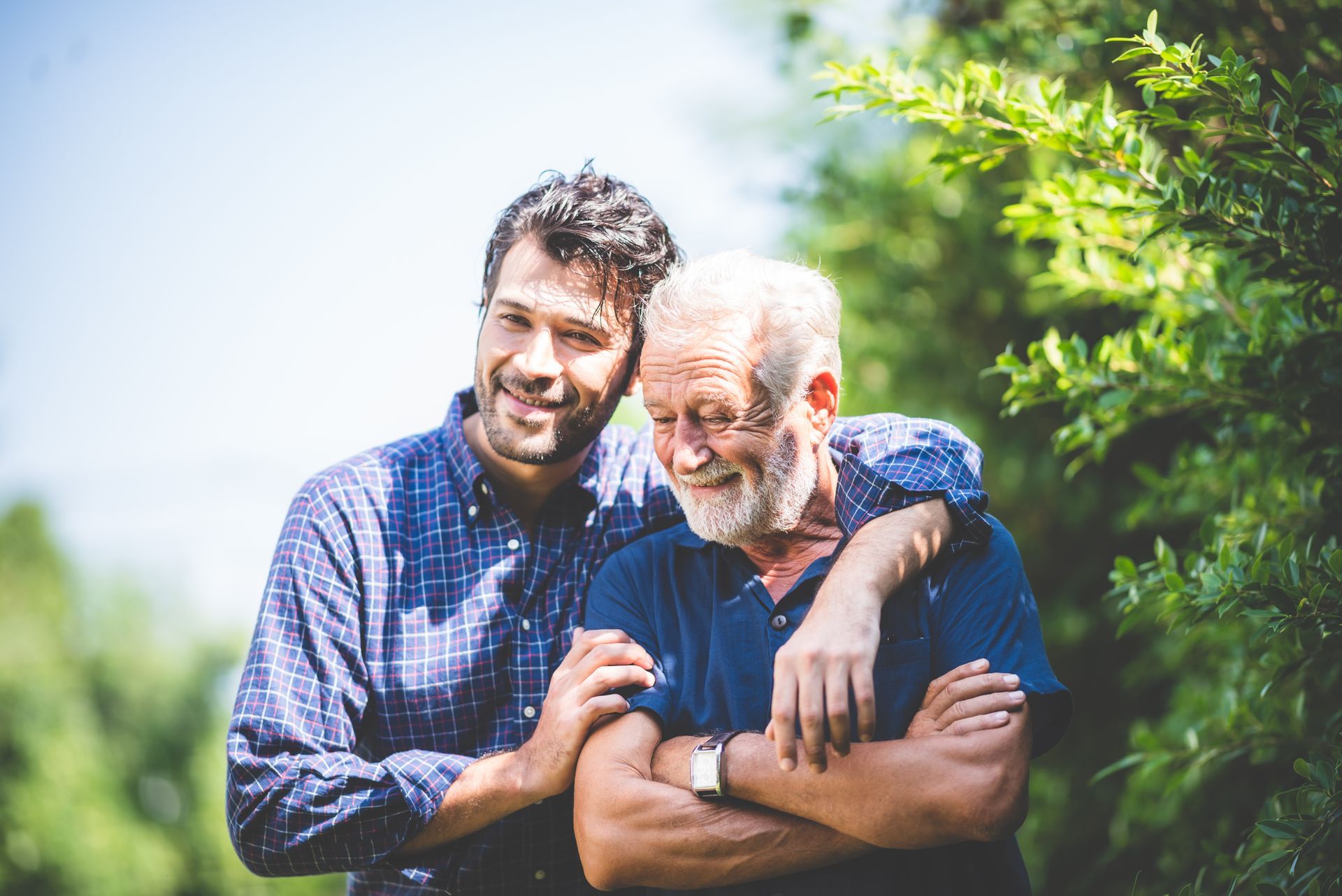 Two smiling people standing outdoors in a green garden, one with an arm around the other.