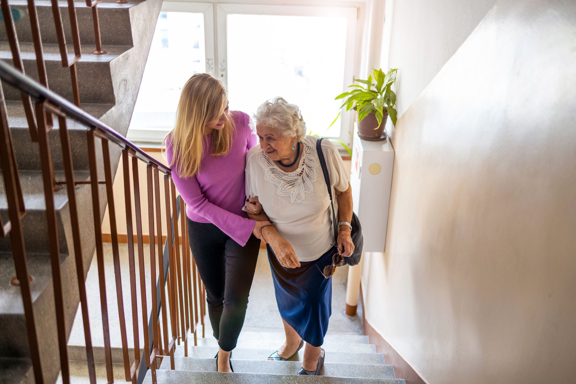 Two women walking up a staircase, one helping the other with a cane, in a bright hallway.
