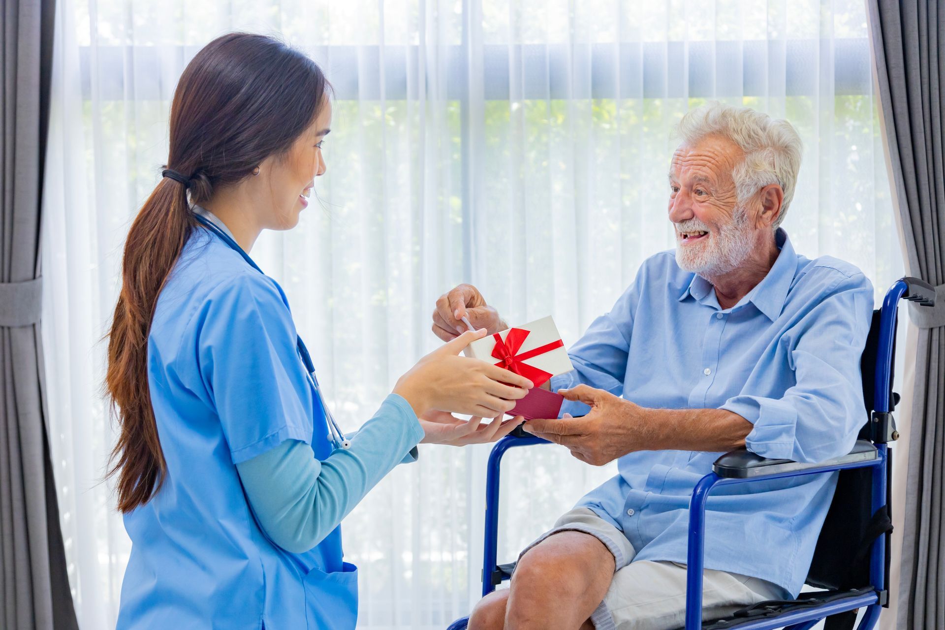 Caregiver giving a wrapped gift to a person in a wheelchair, smiling in a well-lit room.