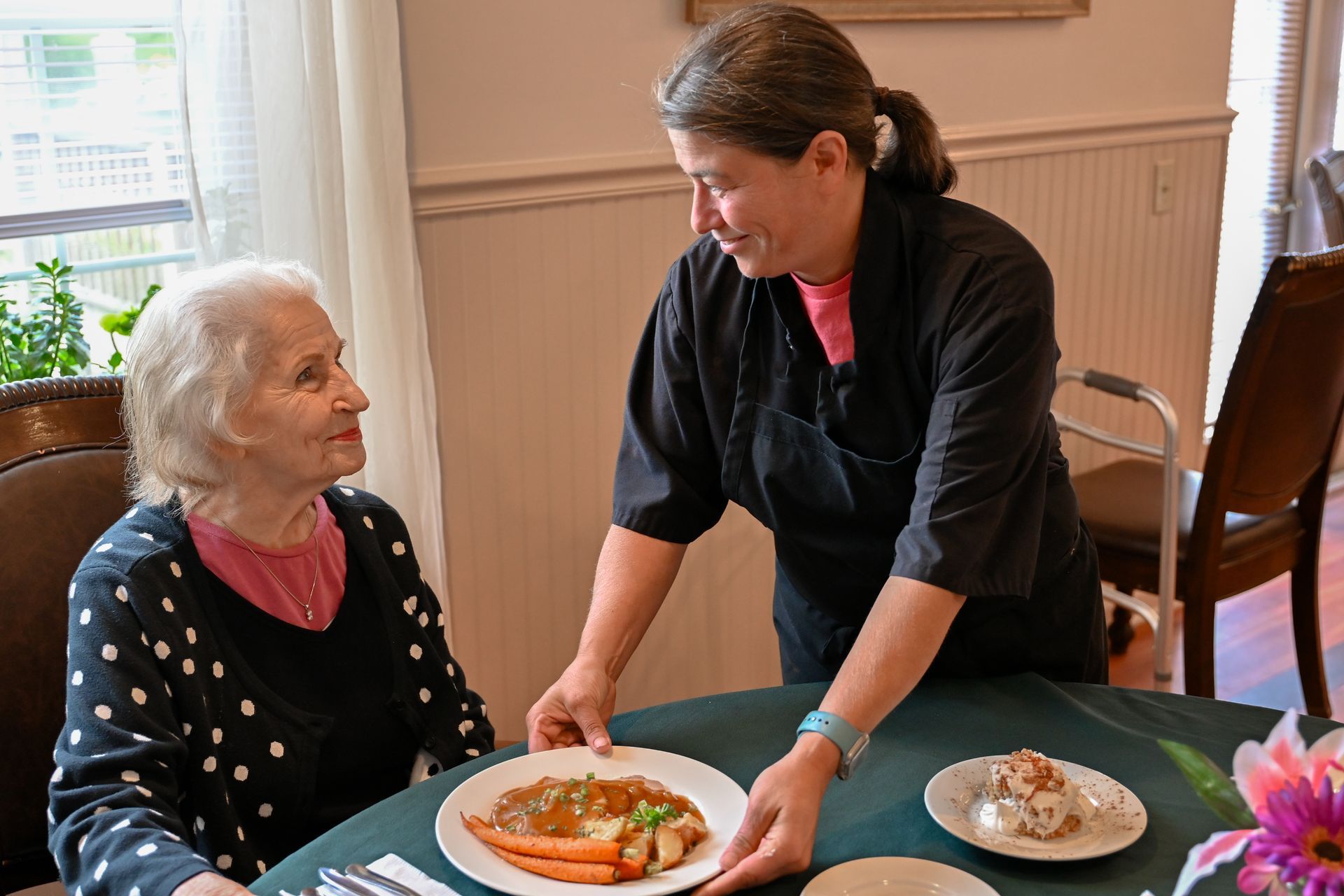 Caregiver serving a plate of food to an elderly person at a table in a dining room.