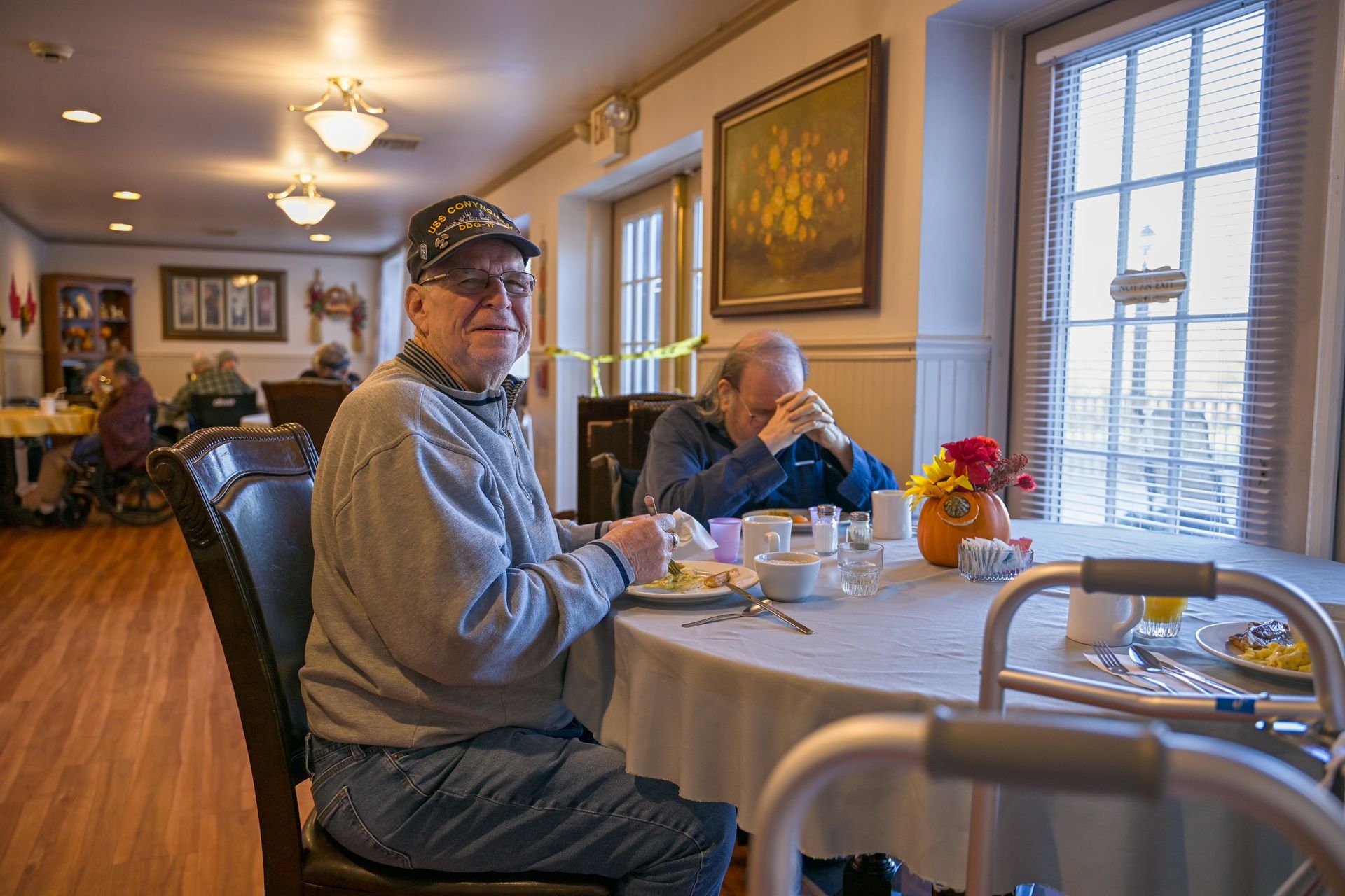 Man at table in a dining room, eating, looking at the camera; another man with head down.
