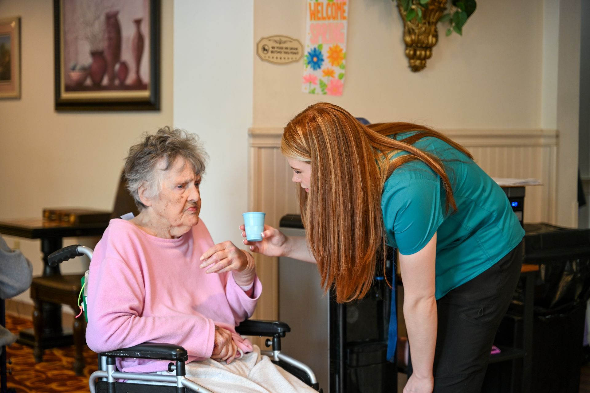 Woman in teal shirt offers drink to elderly person in pink shirt seated in wheelchair.