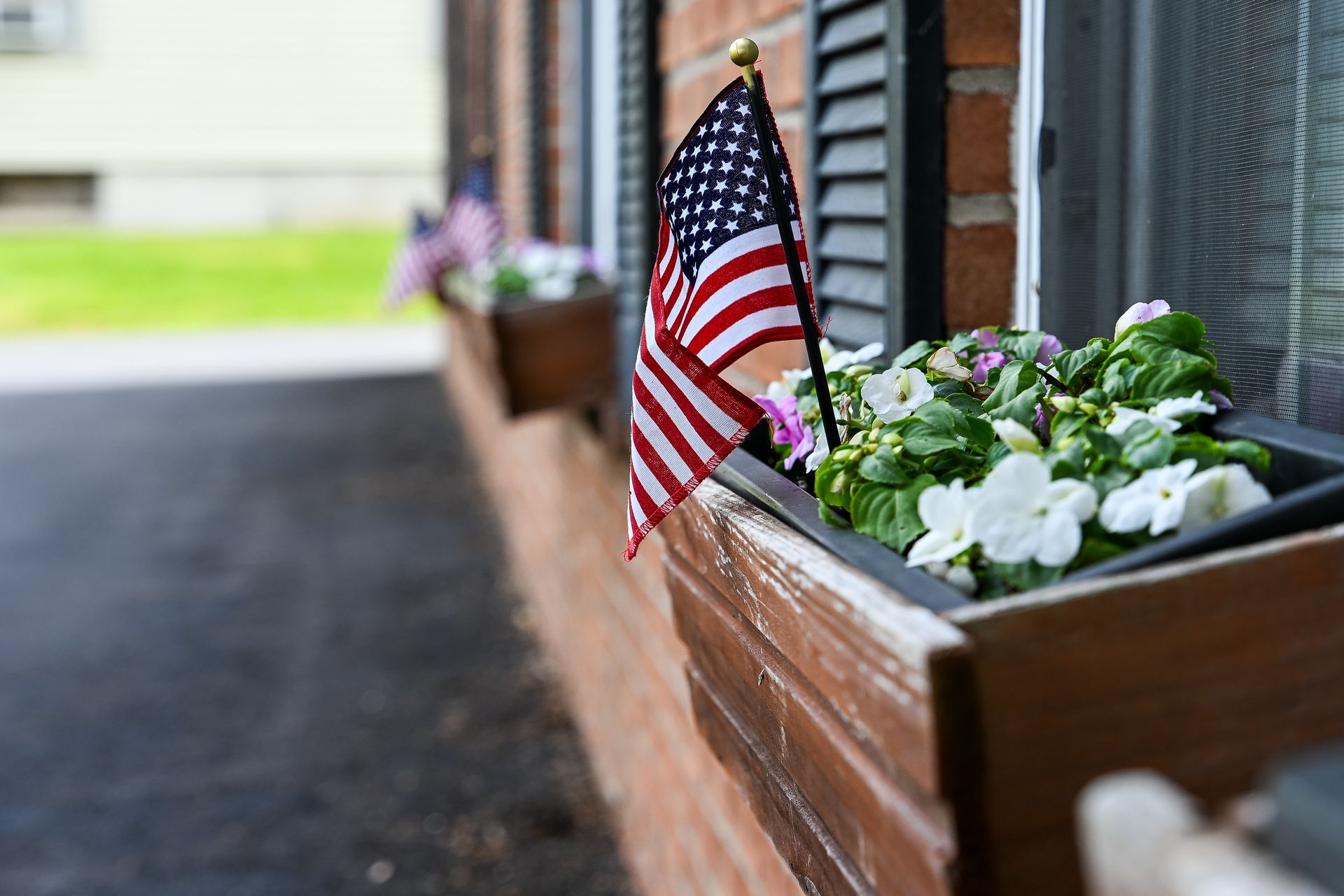 American flag in a wooden planter with white and pink flowers, set against a brick building and window.