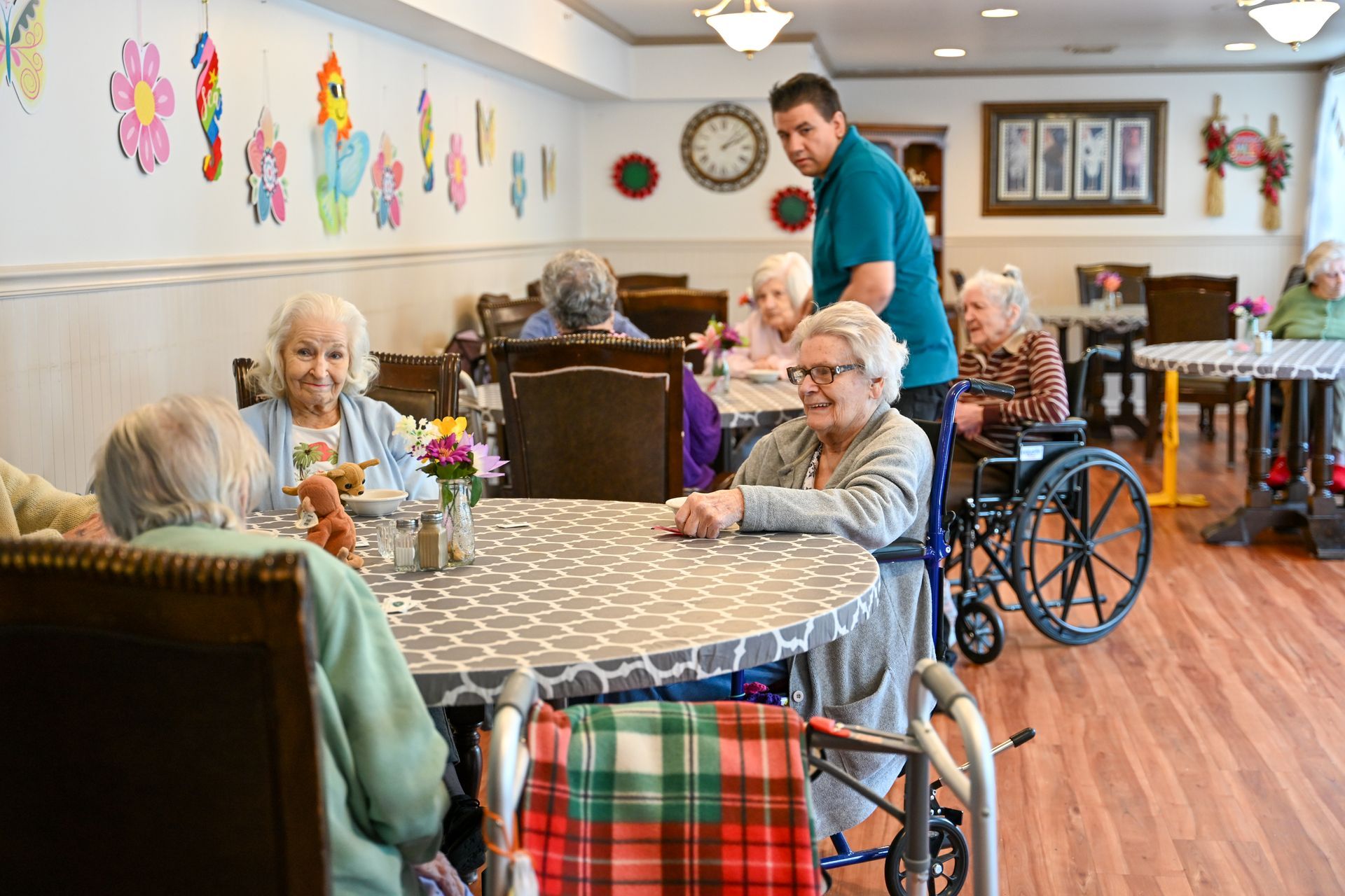 Senior residents seated at tables in a brightly decorated dining room. A caregiver assists one resident.