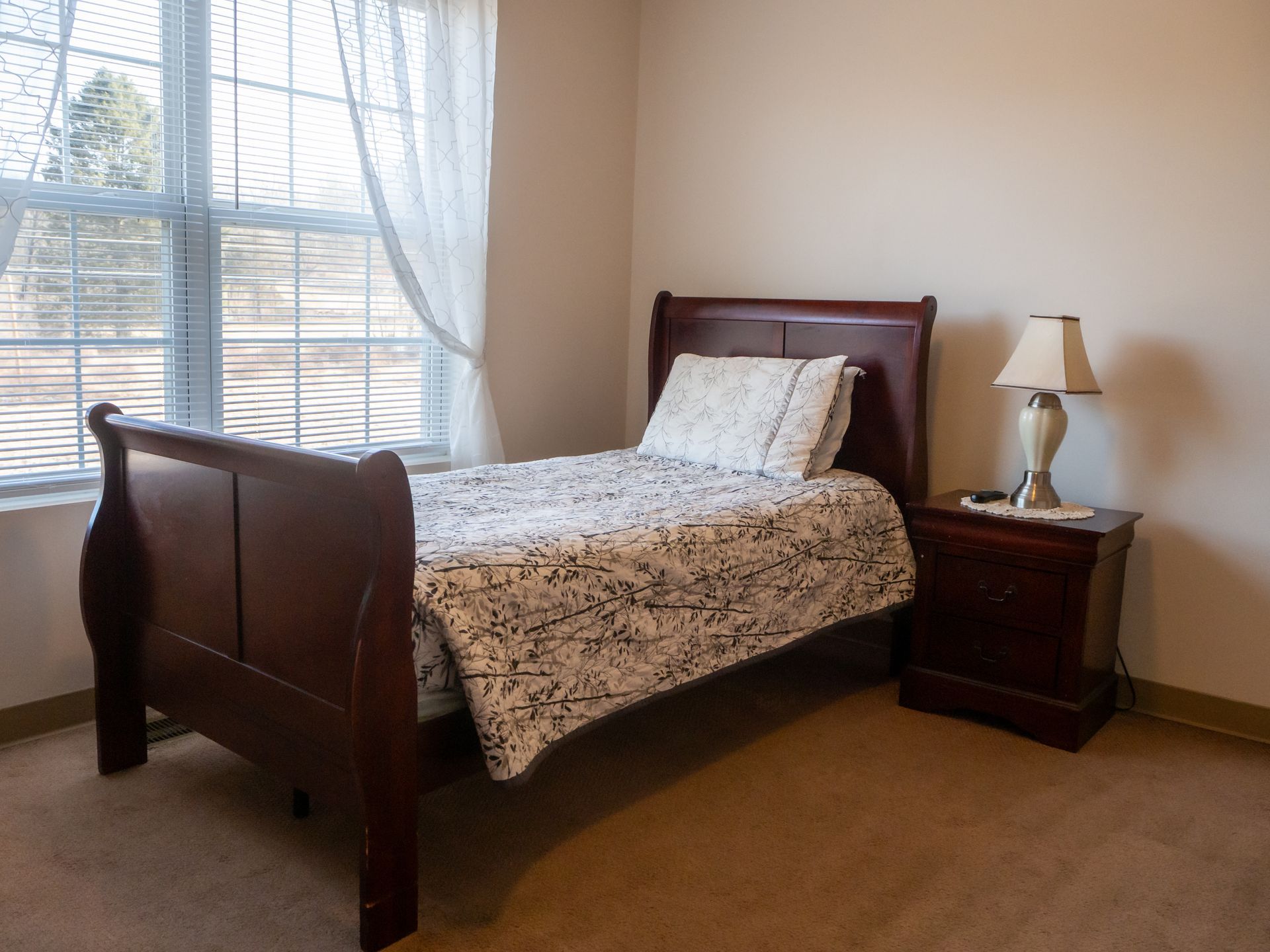 Bedroom with a dark wood bed, nightstand, and window with blinds and sheer curtain.