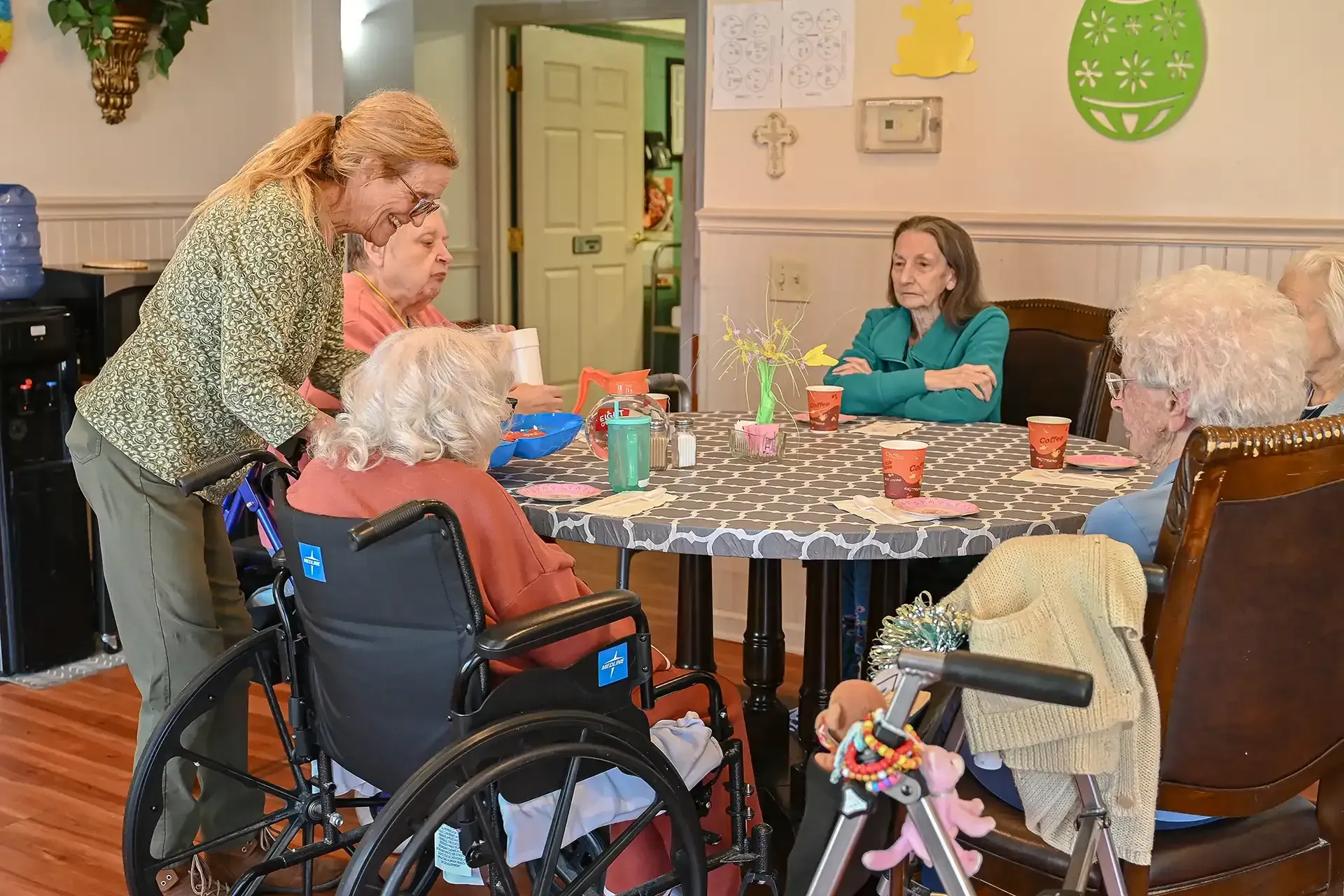 A group of elderly women in a nursing home sit at a table, being assisted by a caregiver.
