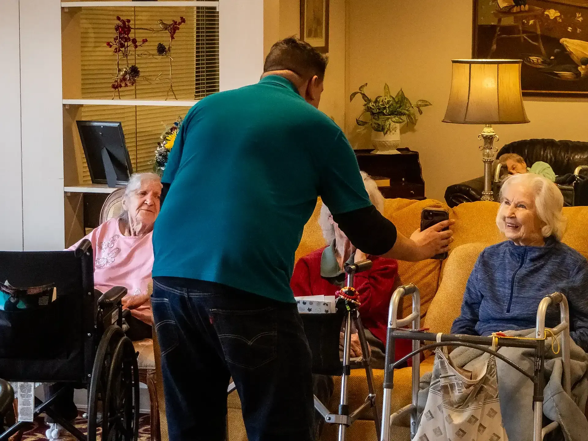 Man in teal shirt takes a photo with three older women in a living room, two in wheelchairs.