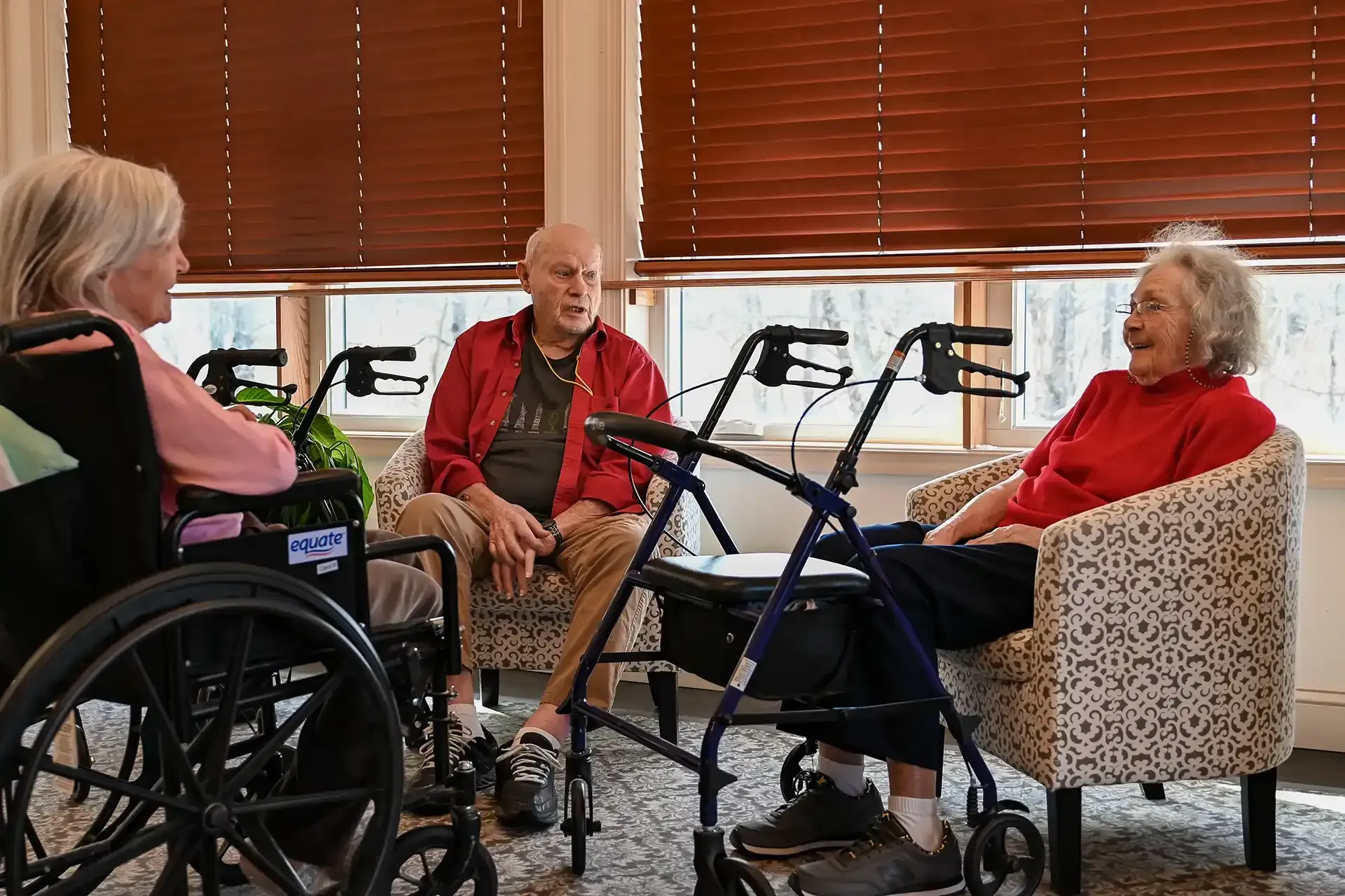 Three older adults chatting in a room with sunlight; one in a wheelchair, two in armchairs with walkers nearby.