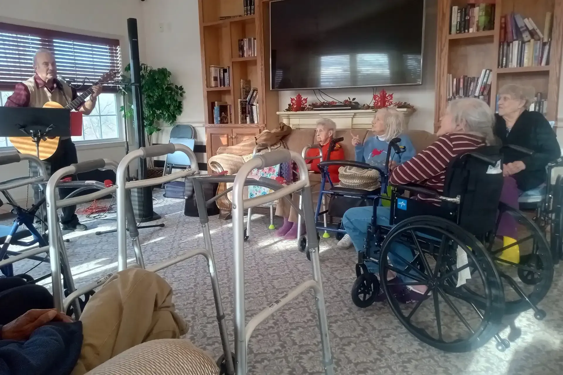 A musician plays guitar for a group of seniors in a living room. Some are in wheelchairs, enjoying the music.