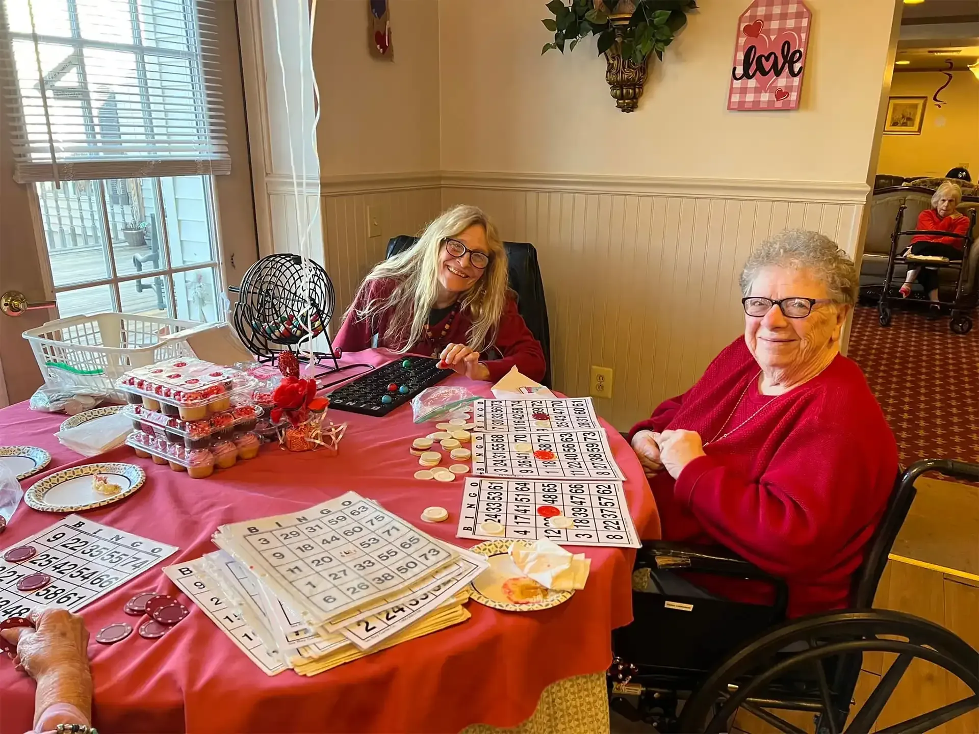 Two women playing a game at a table. One is in a wheelchair, wearing red. The other smiles. Valentine decor.