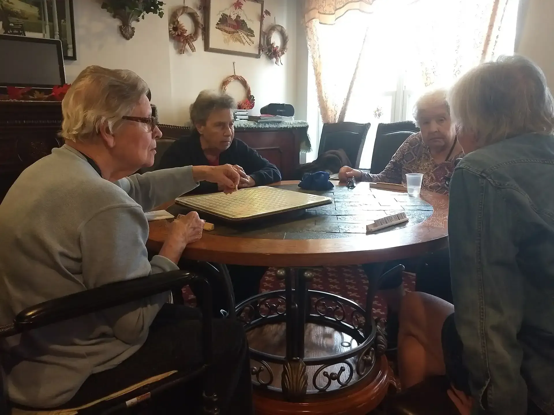 Four elderly women playing a board game around a round table indoors.