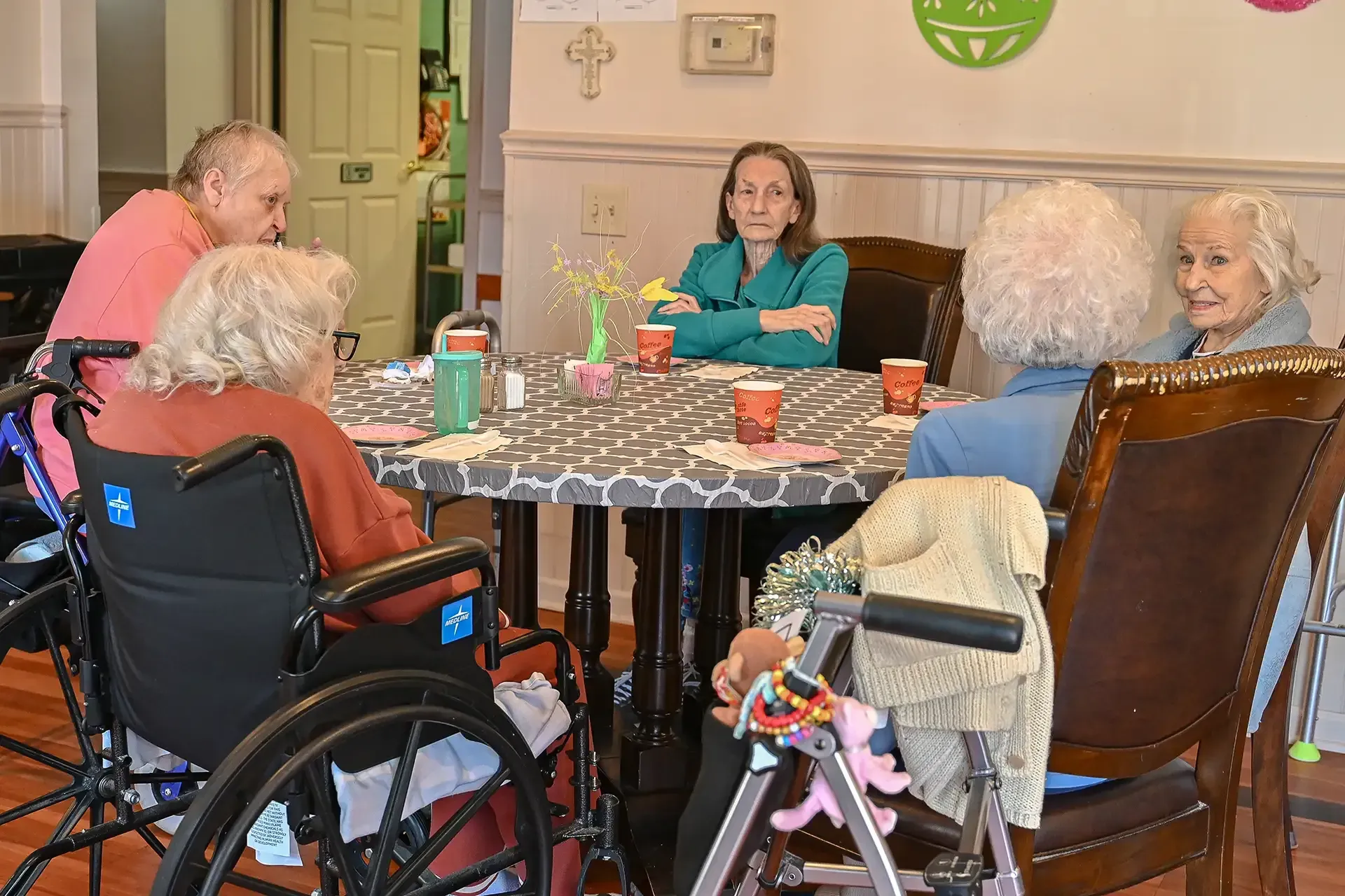 Group of seniors and woman at a table, some in wheelchairs, smiling and chatting. Indoors.
