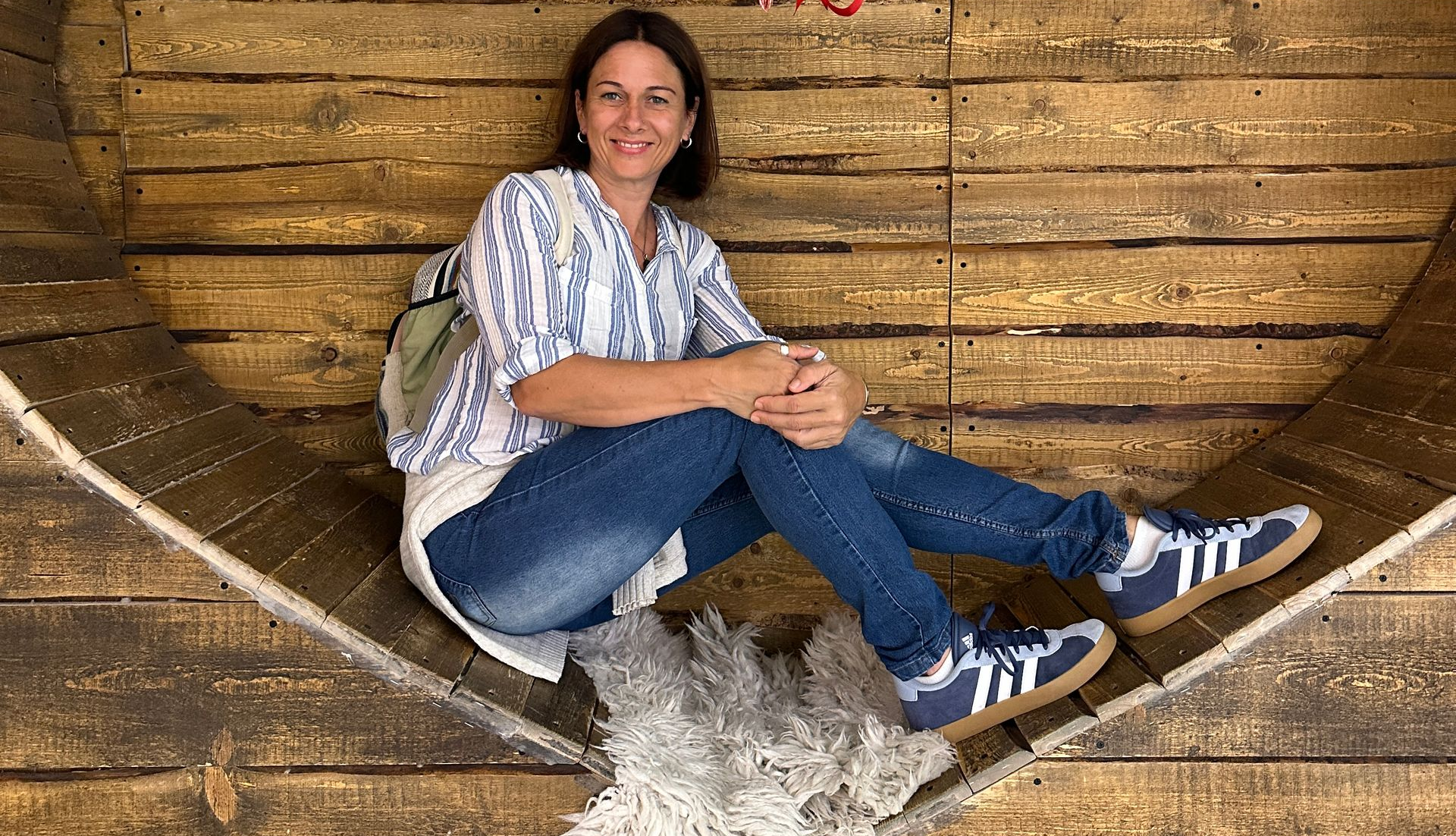 Frau sitzt im Schneidersitz auf einer Holzkonstruktion. Sie trägt Jeans, gestreiftes Hemd und Turnschuhe. Im Hintergrund sind Holzbretter.