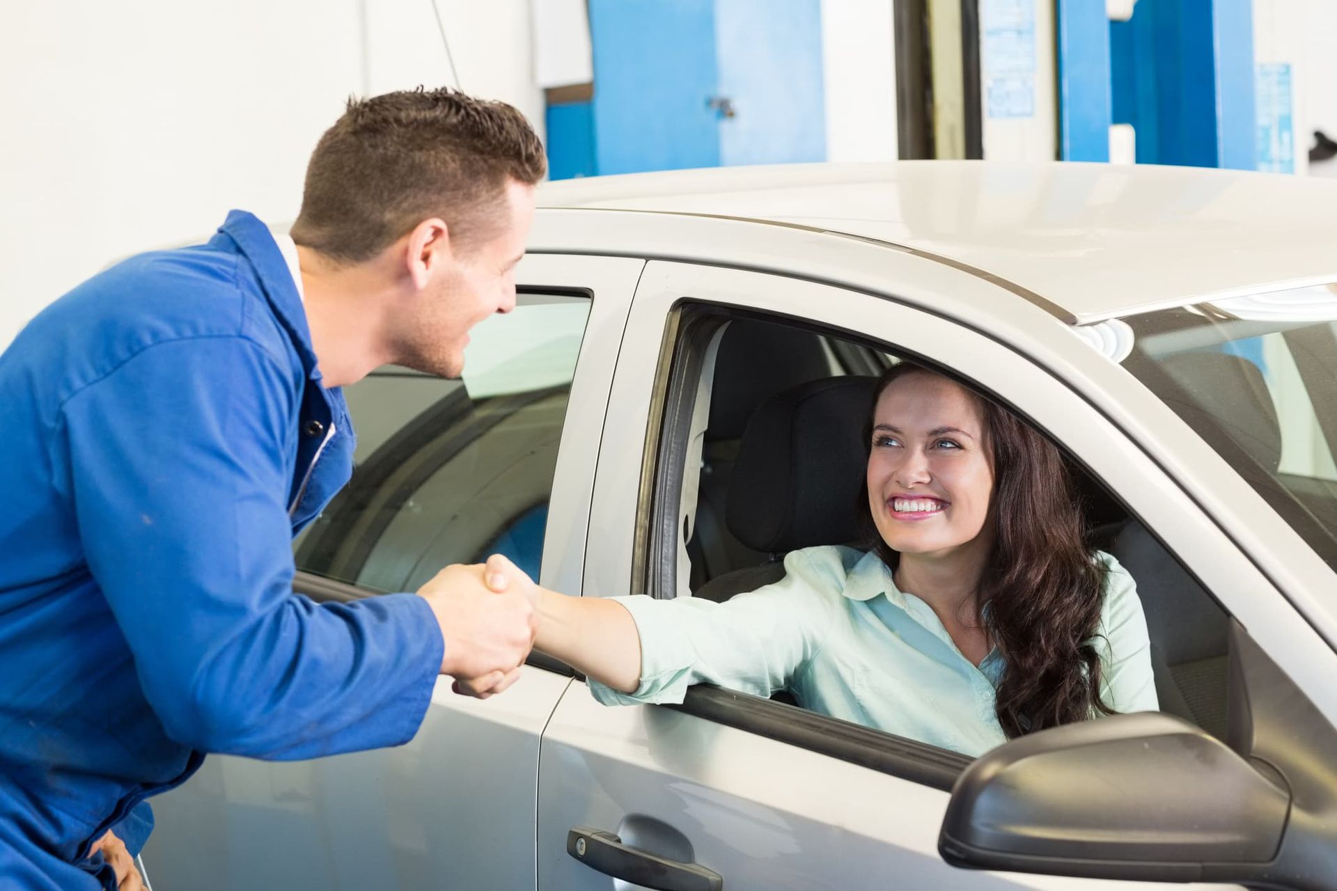 Mechanic shakes hands with a smiling woman in a silver car at a garage.