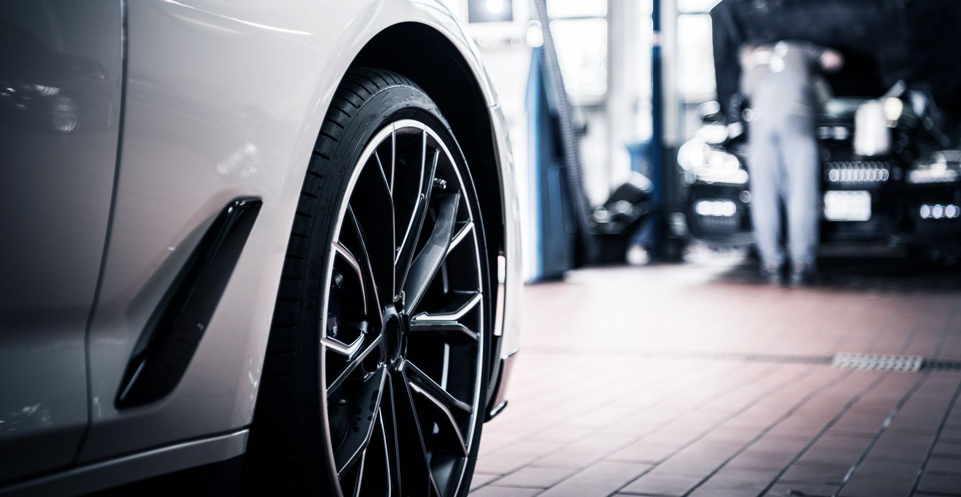 Silver car in a repair shop with a black tire and rim; mechanic in the background.