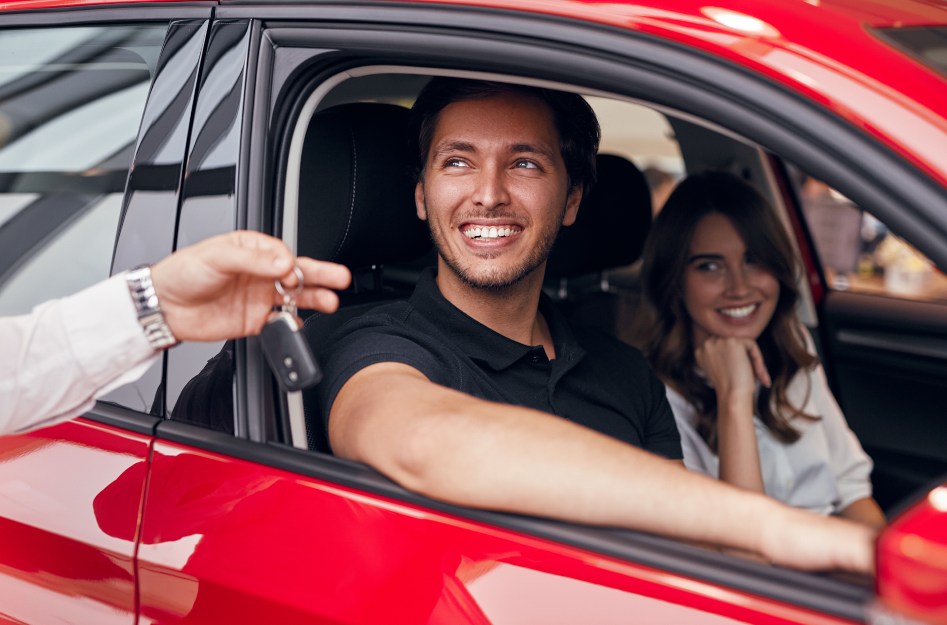 Man and woman smiling in red car, receiving keys from hand.