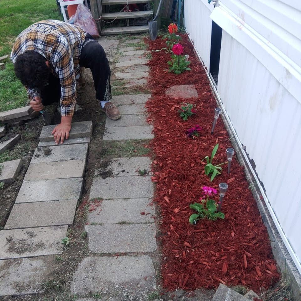 Person installing paver walkway beside a fresh red-mulch flower bed with small pink flowers