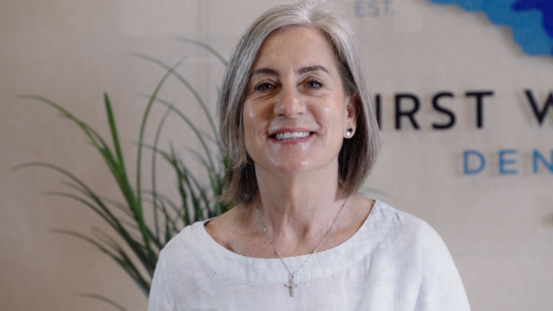 Woman smiling in a dental office, in front of a sign and plant.