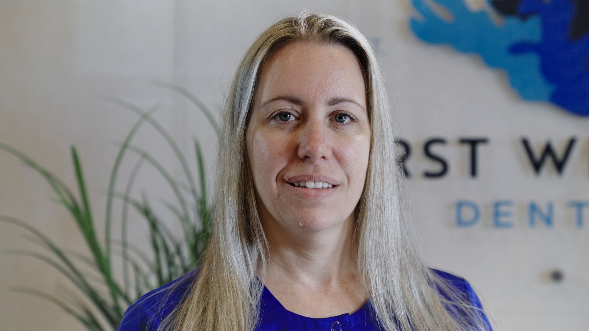 Woman with long blonde hair, wearing a blue shirt, smiling in a dental office.