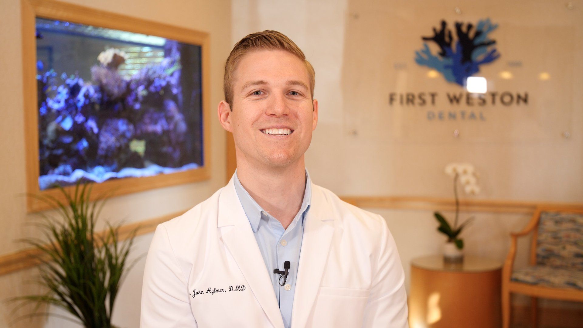 Man in white coat smiles, standing in a dental office. Fish tank and logo visible in the background.