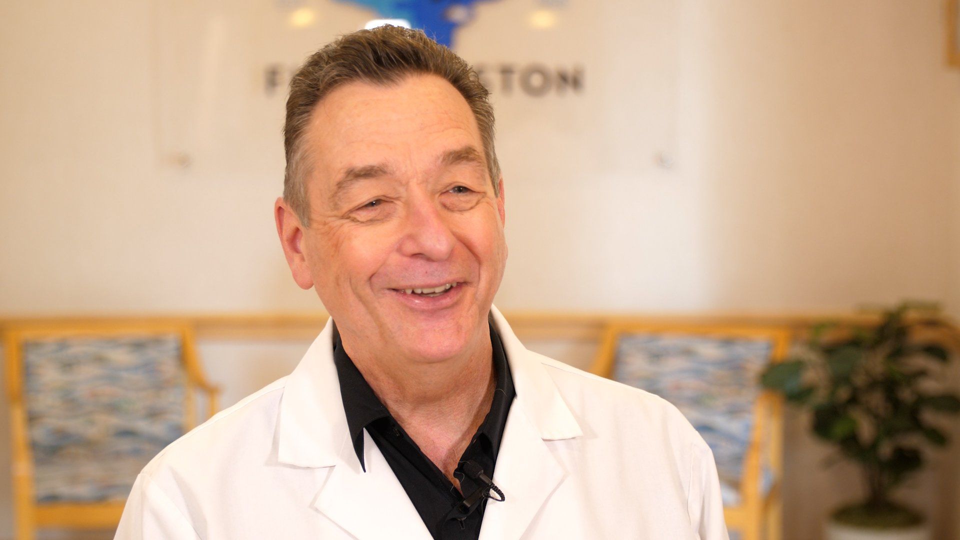 Smiling man wearing a white lab coat, indoors in an office setting.