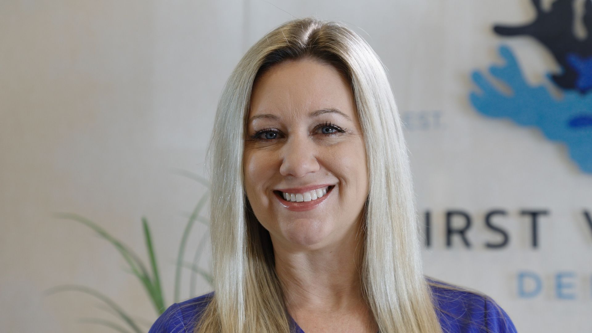 Woman with long blonde hair smiles, standing in an office setting with text and a blue graphic on the wall behind her.