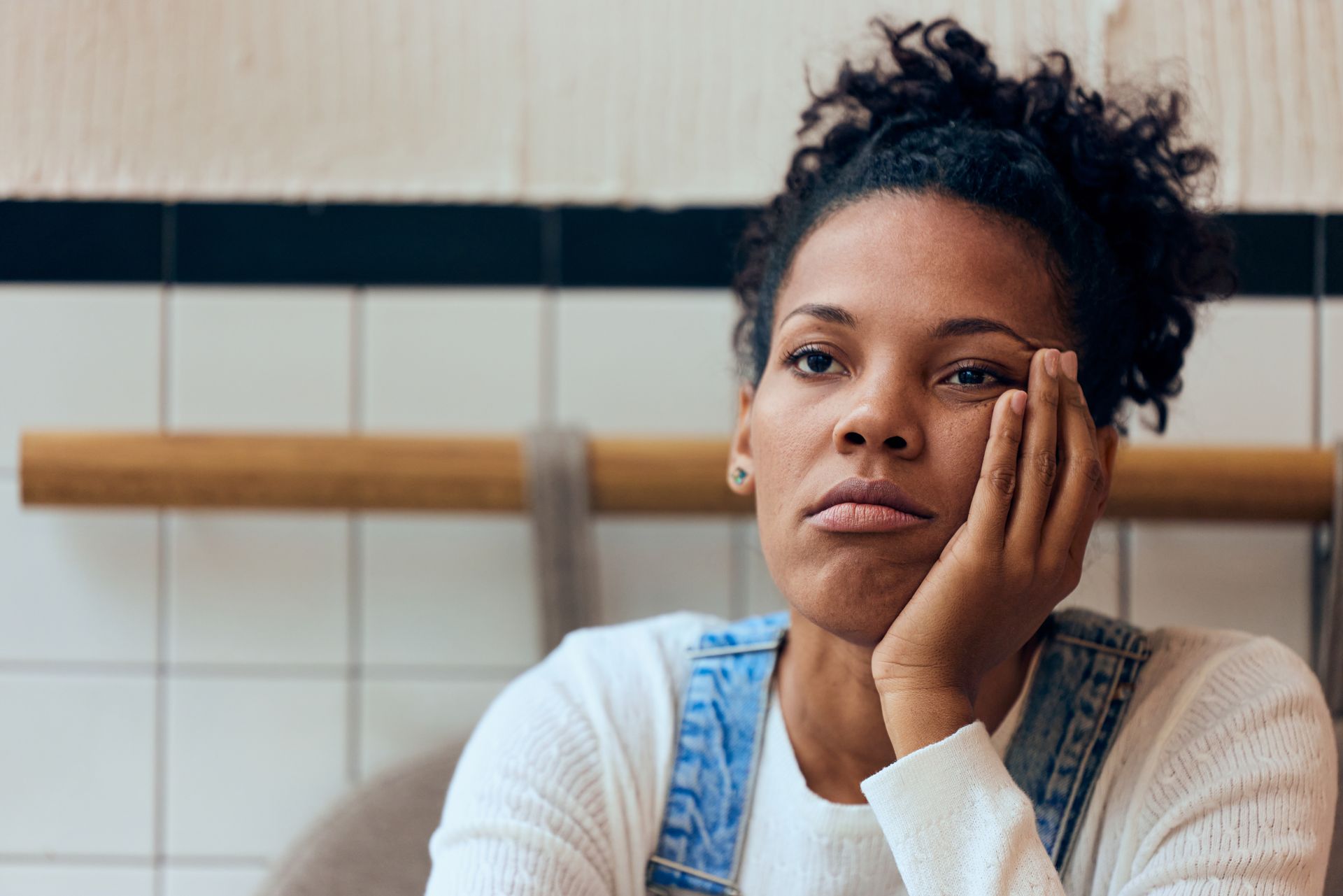 Woman resting head on hand, looking bored. White top and denim overalls; tiles in background.