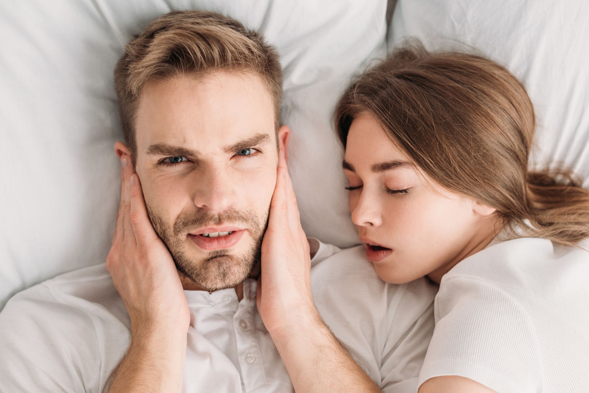 Man covering ears in bed next to a woman sleeping and snoring.
