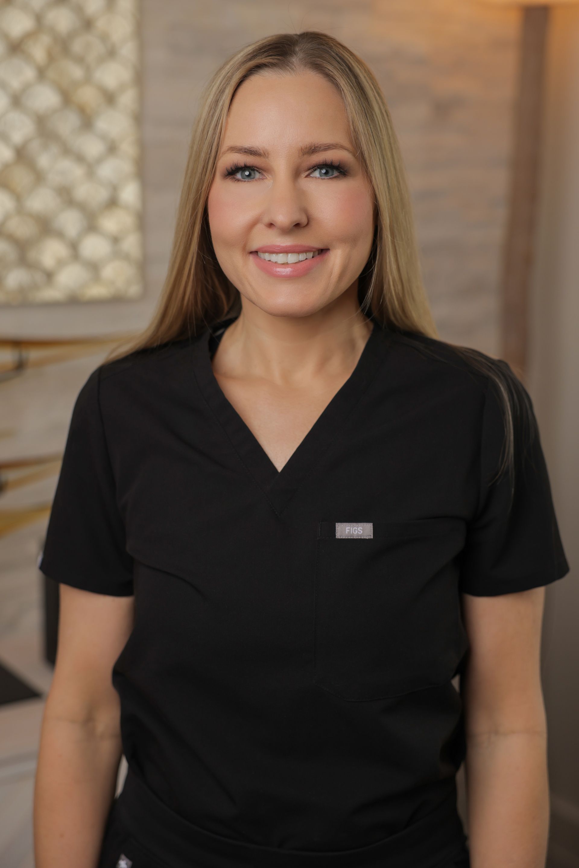 A woman in a black scrub top is standing in a room and smiling.