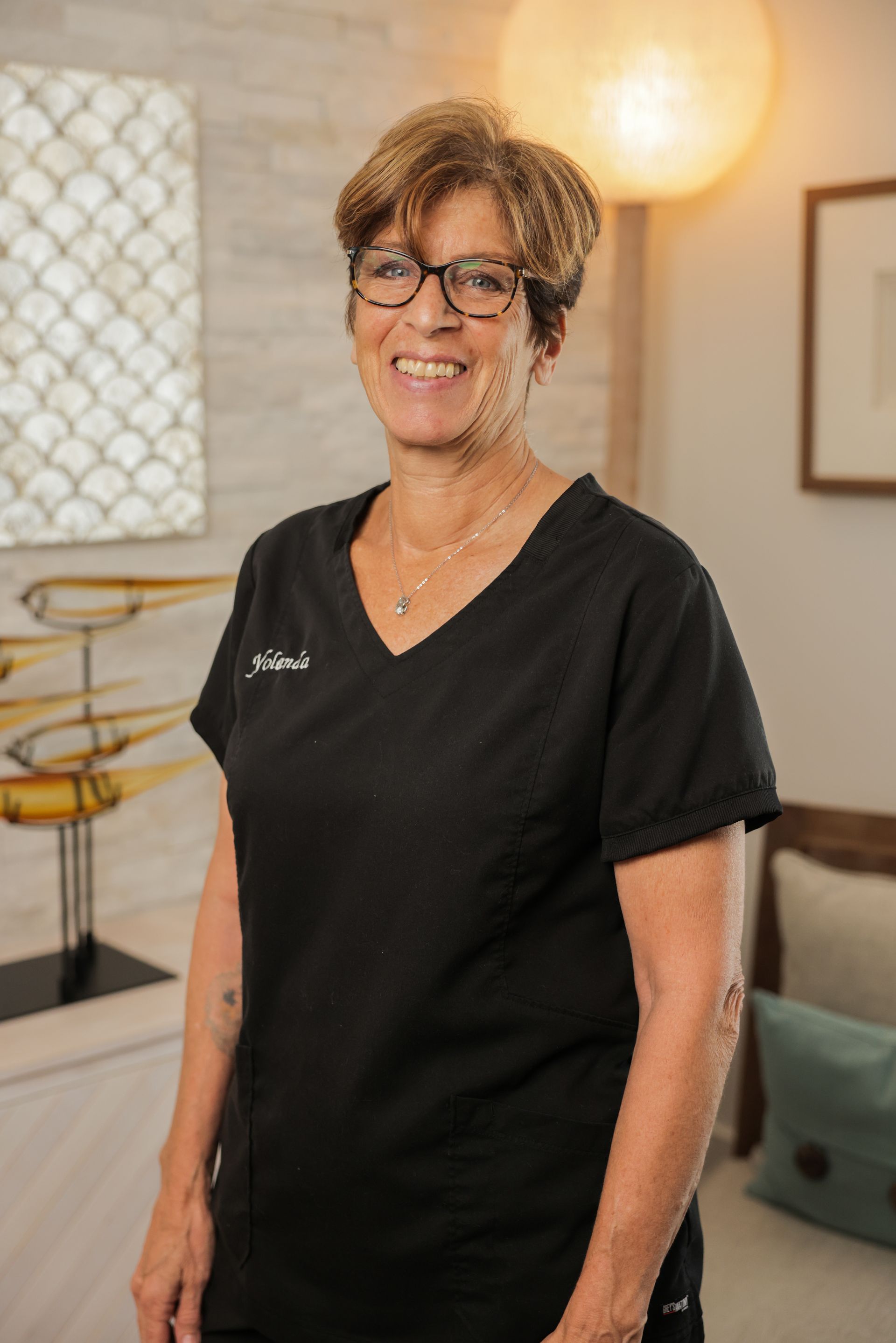 A woman in a black scrub top and glasses is standing in a living room.