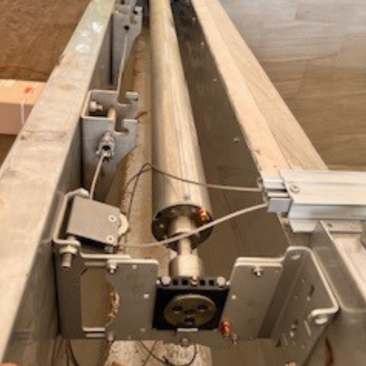 Silver cylindrical component in a metal frame, likely machinery. Wires and mounting brackets visible.