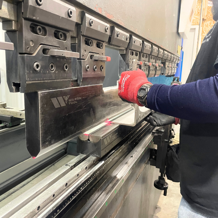 Person using a press brake machine to bend metal. Red gloved hand manipulates metal sheet. Industrial setting.