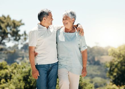 Two people walking side by side outdoors, smiling at each other with their arms around one another.