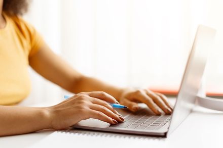 A person in a yellow shirt sits at a desk using a silver laptop while holding a blue pen.