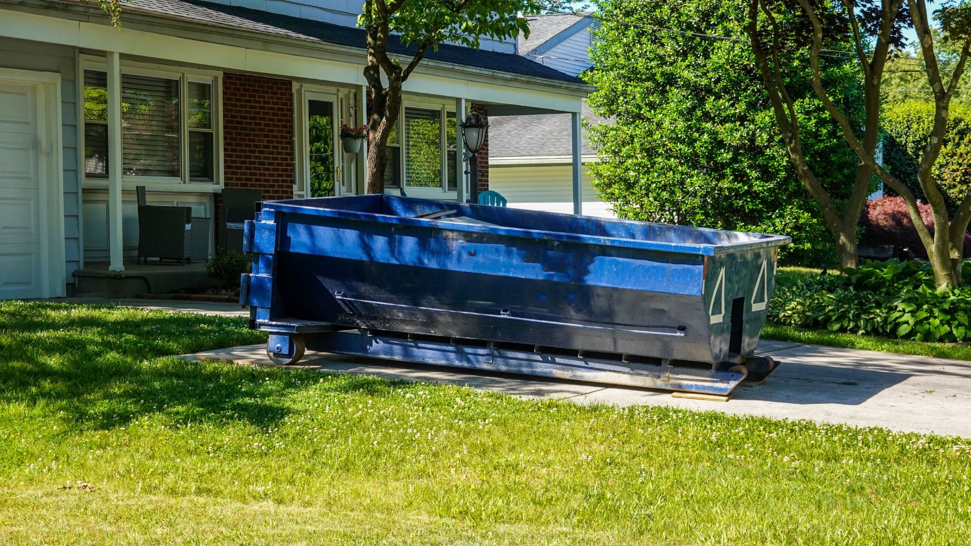 A blue dumpster is sitting in the grass in front of a house.