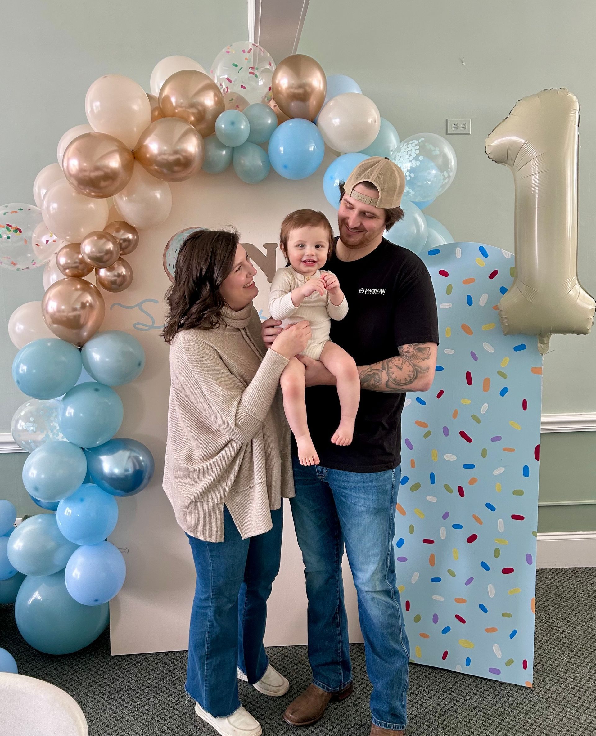 A man and woman are holding a baby in front of a balloon arch.