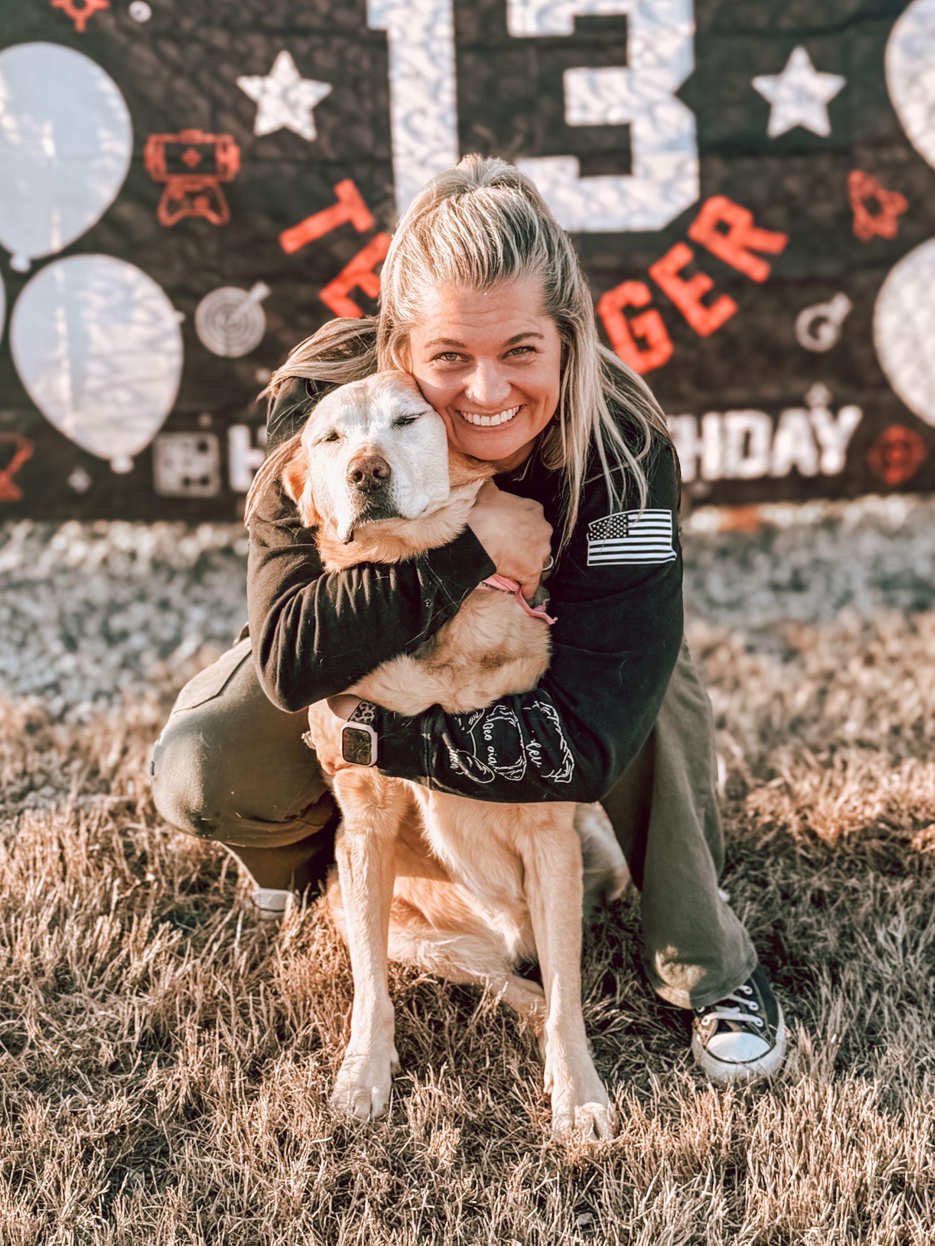 Bonnie is kneeling down, holding a dog in front of a birthday banner.