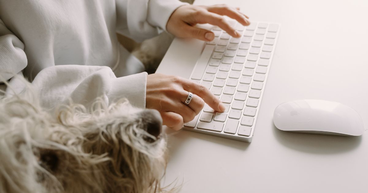 A person is typing on a keyboard next to a dog.
