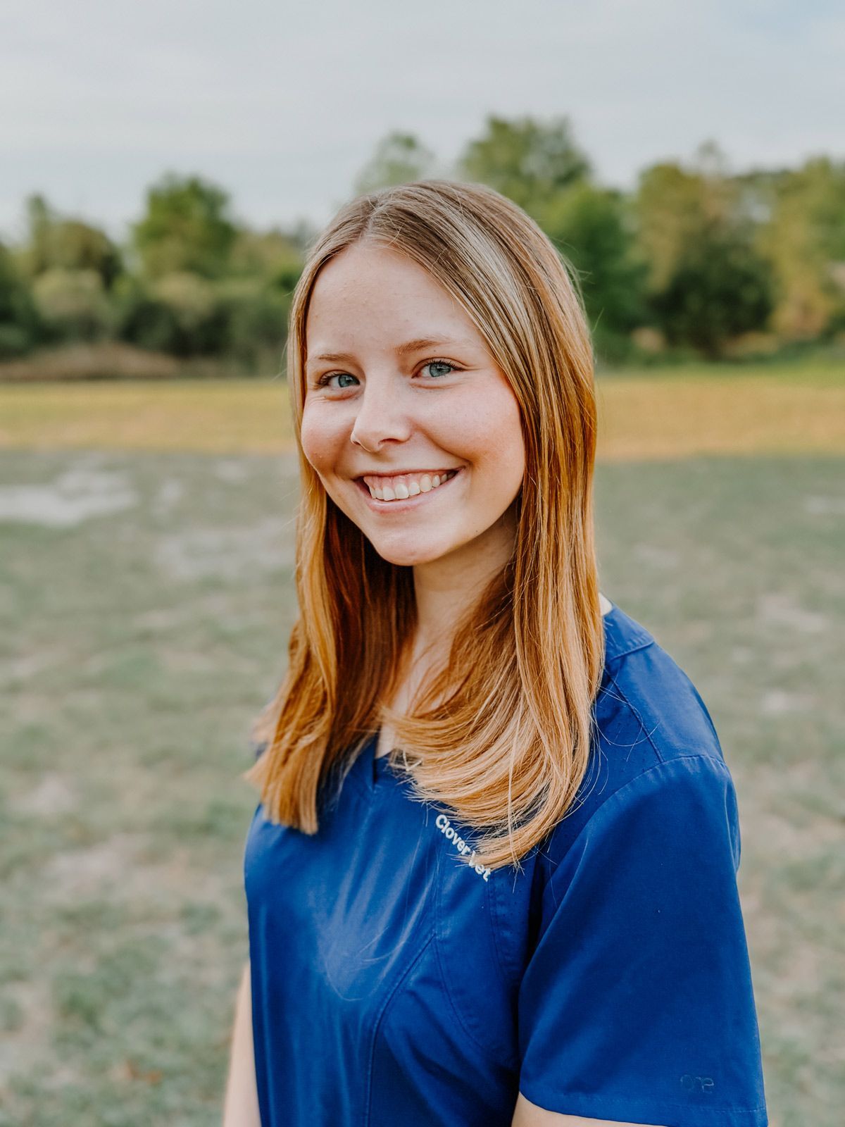 A woman in a blue shirt is smiling for the camera while standing in a field.