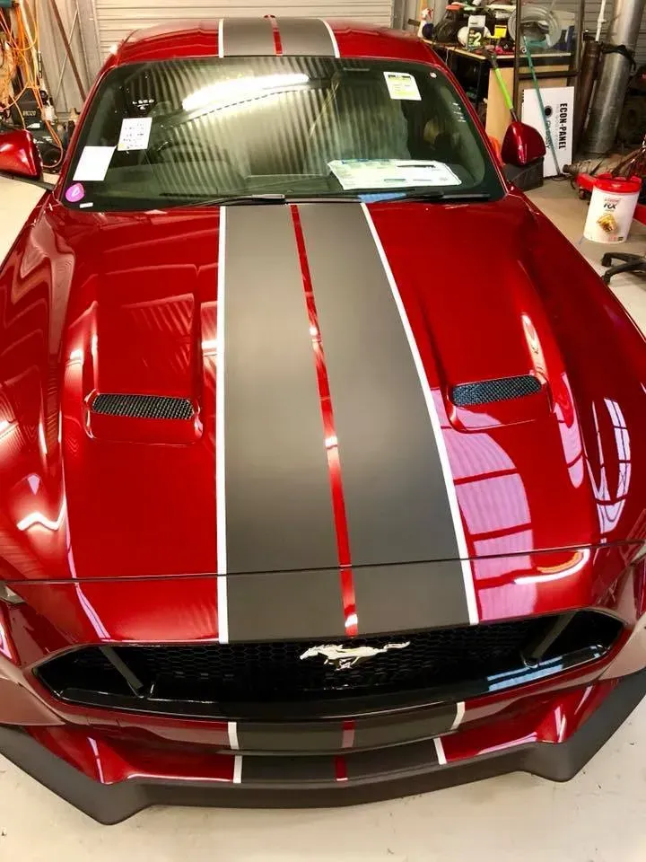 A red mustang with black and white stripes on the hood is parked in a garage.