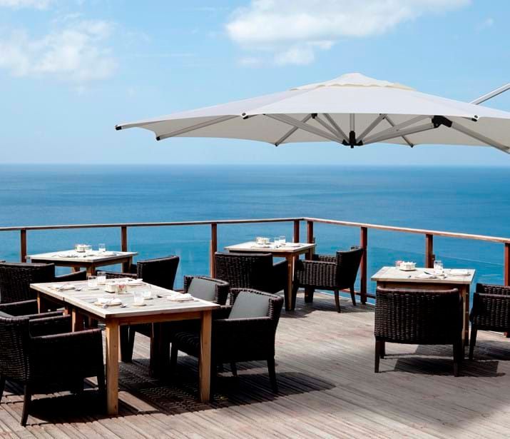 Outdoor dining area overlooking the ocean with tables, chairs, and a large umbrella. Outdoor dining area overlooking the ocean with tables, chairs, and a large umbrella.