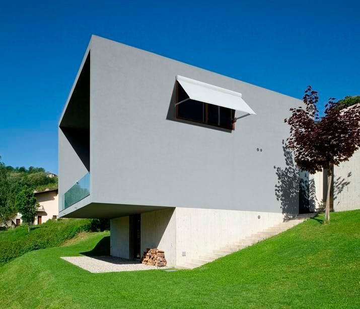 Modern gray house with angled roof, cantilevered balcony, and awning over a window on a grassy hillside.