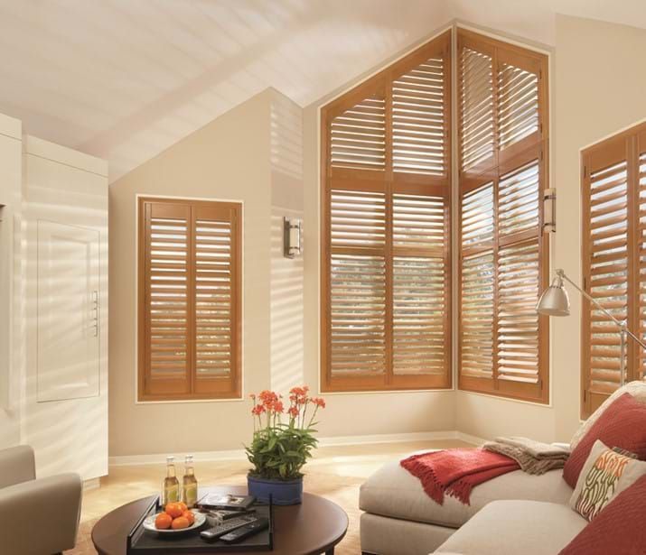 Living room with wooden shutters on various windows, including a large triangular window, soft lighting.