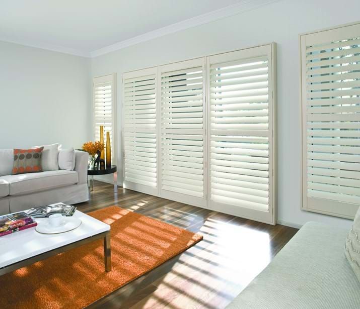 Living room with white shutters, couch, orange rug, and coffee table. Sunlight streams through.