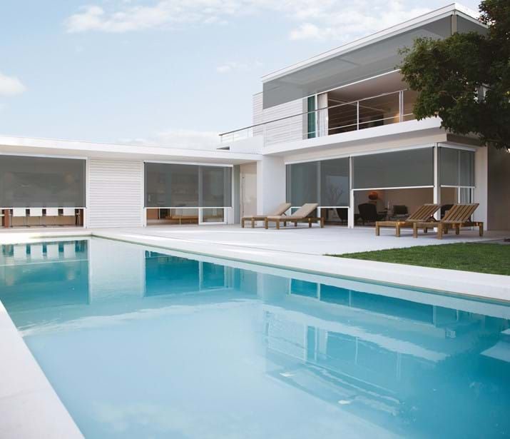 Modern home with a pool, shaded windows, and lounge chairs. Blue pool, white house, sunny day.